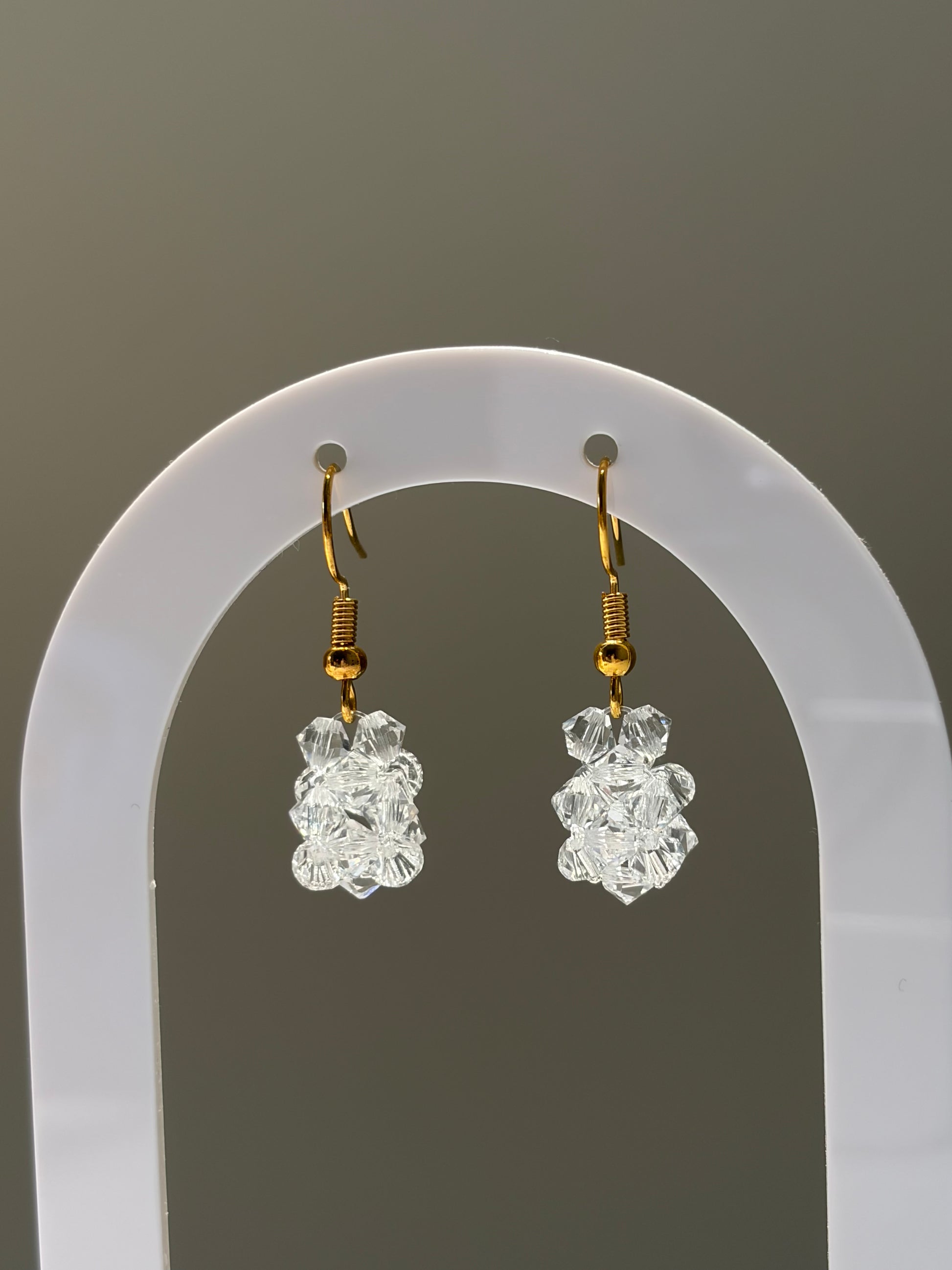 A pair of gold-tone dangle earrings with clear faceted Swarovski crystal beads, displayed on a white arched jewelry stand against a neutral gray background.