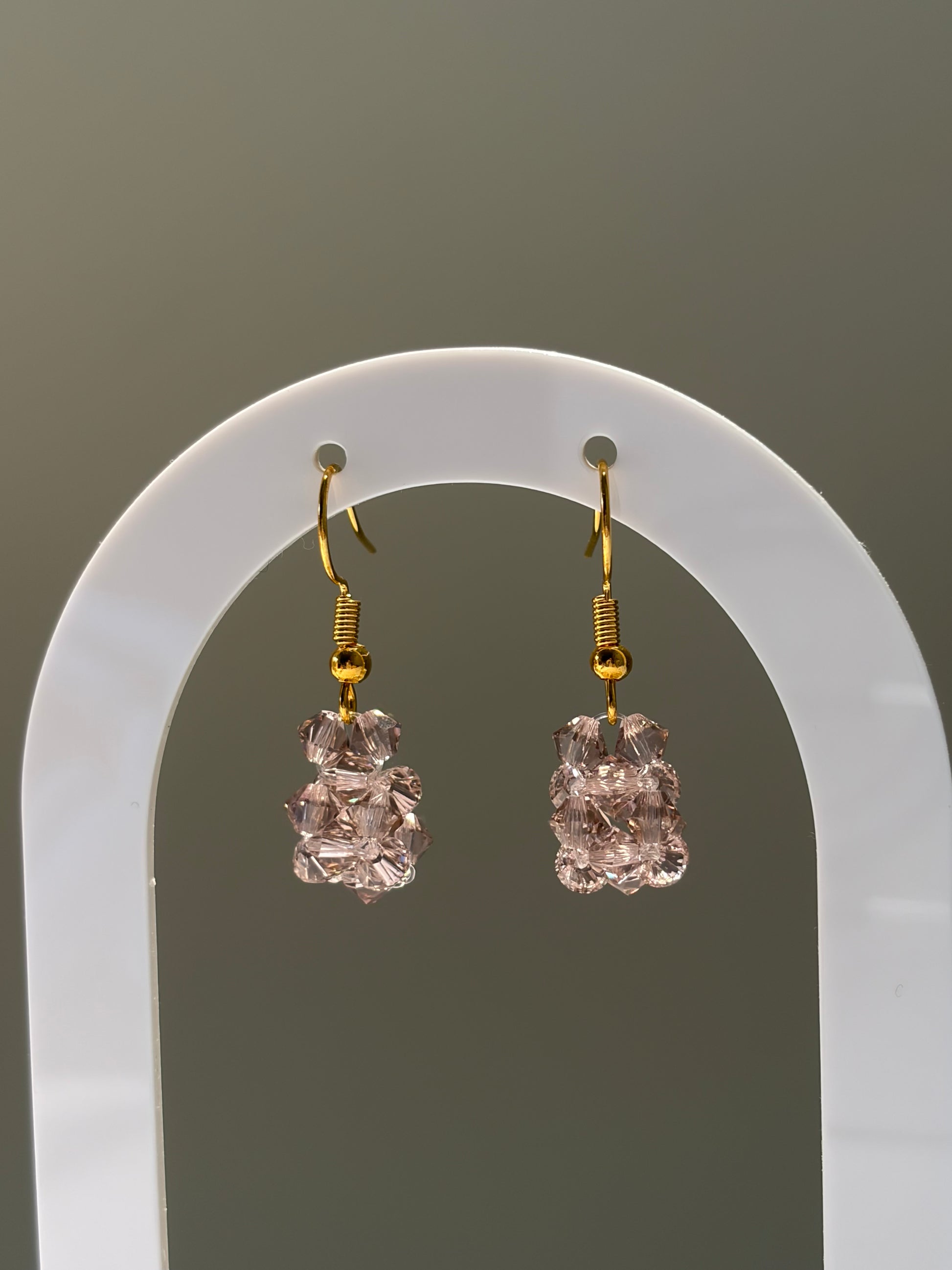 A pair of gold-tone solid dangle earrings with faceted rose gold Swarovski crystal beaded drops, displayed on a white arched stand against a neutral gray background.