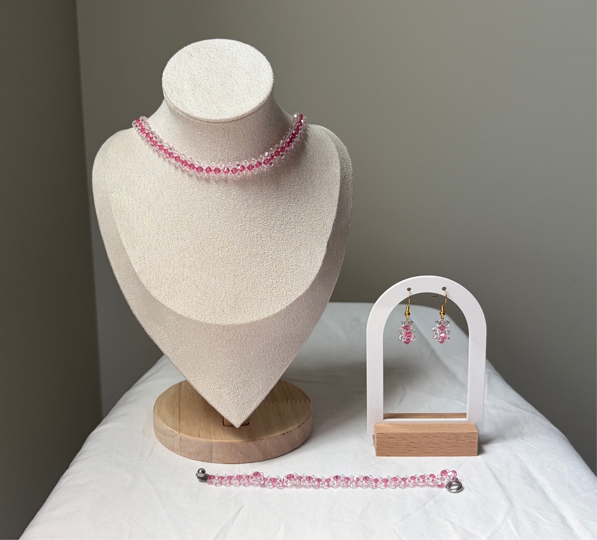 A Swarovski crystal beaded jewelry set displayed on a cream fabric mannequin bust and a small white arch stand, featuring a necklace, bracelet, and matching earrings with pink and white striped design. 