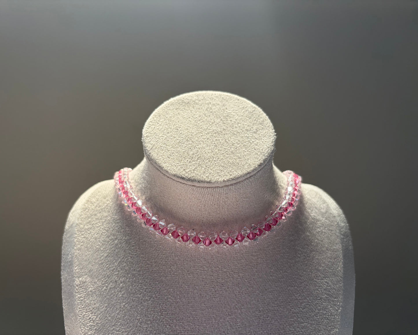 A delicate striped design of pink and clear faceted Swarovski crystal beads draped around a white jewelry display bust against a dark gray gradient background.