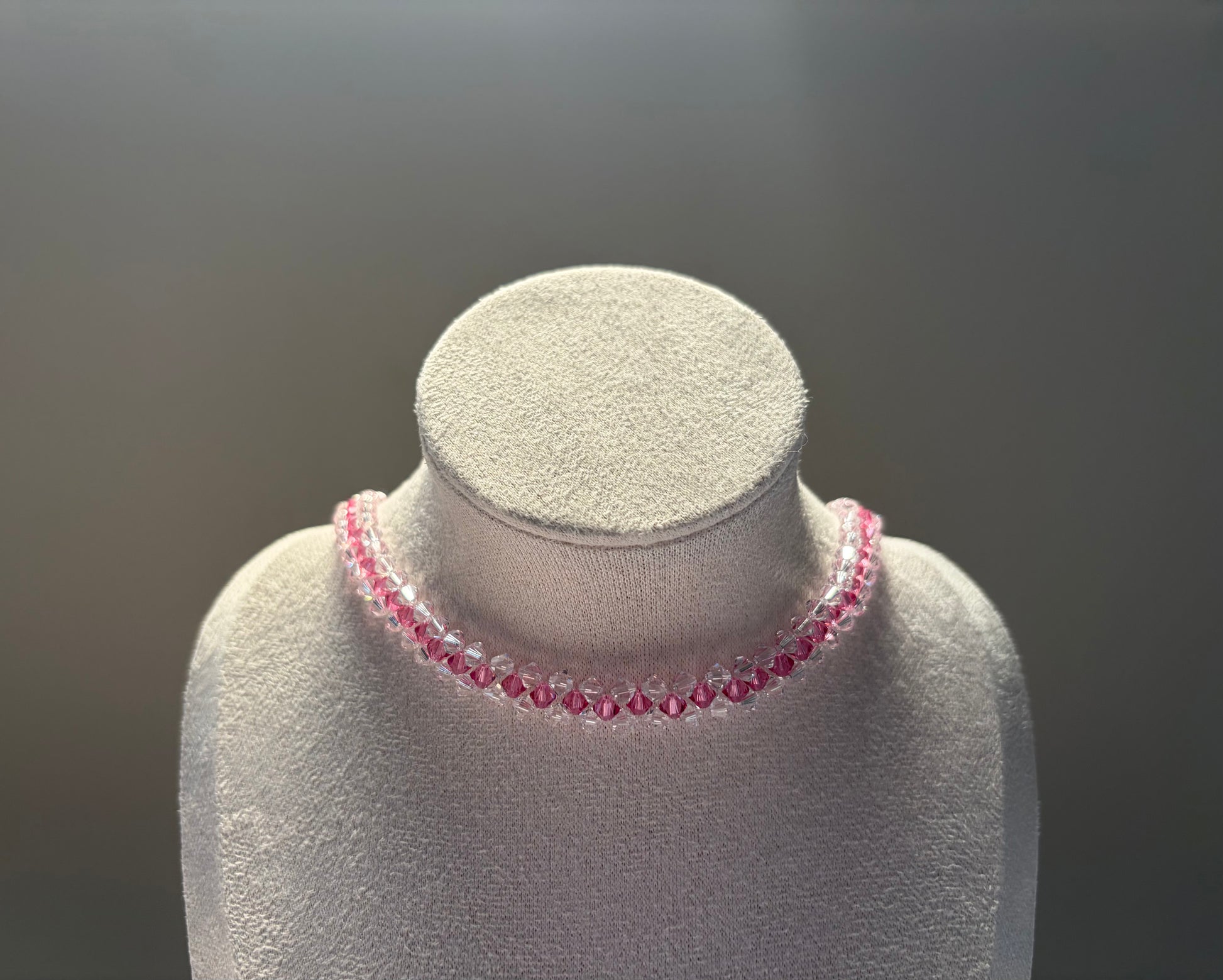 A delicate striped design of pink and clear faceted Swarovski crystal beads draped around a white jewelry display bust against a dark gray gradient background.