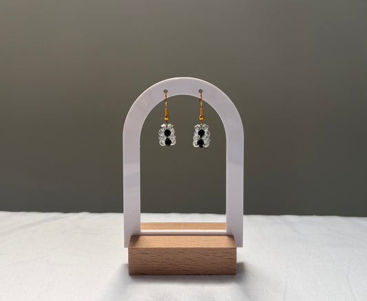 A pair of dangling earrings displayed on a white arch stand with a wooden base. The earrings have gold-toned posts with clear faceted Swarovski crystal beads and black center accents, set against a neutral gray background.