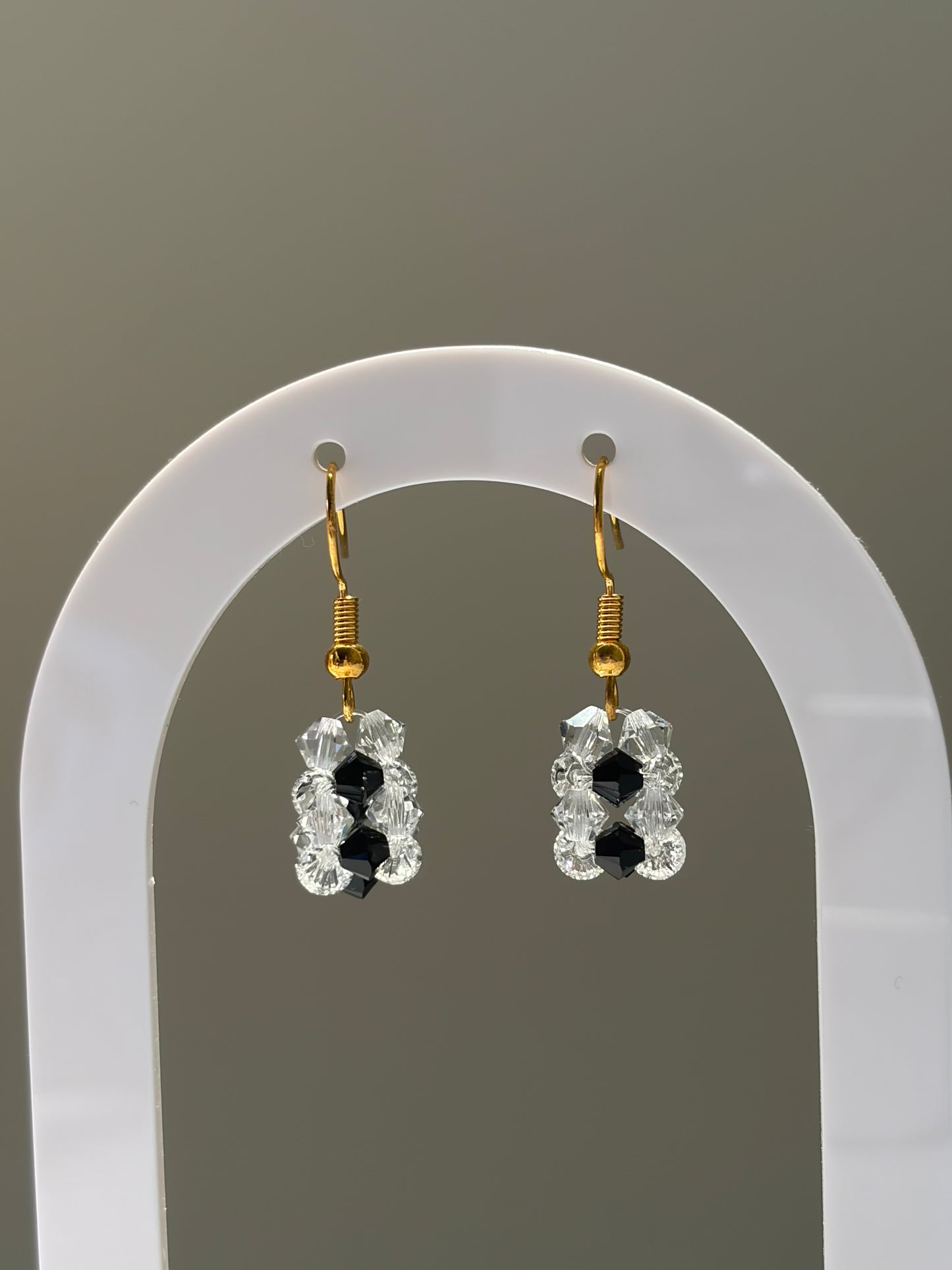 A pair of gold-tone dangle earrings with clear faceted Swarovski crystal beads and small black center beads, displayed on a white arched stand against a neutral gray background.