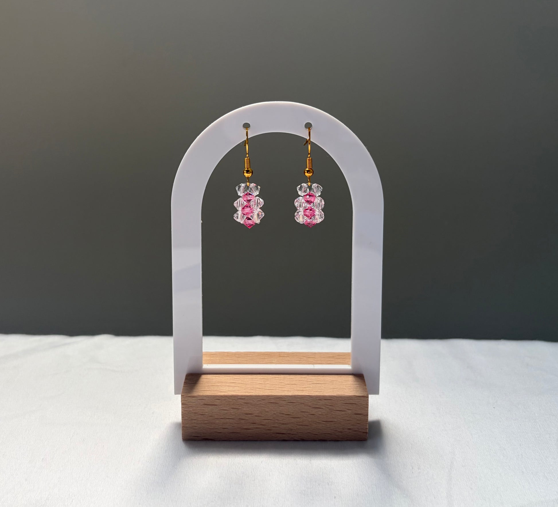 A pair of dangling earrings displayed on a white arched stand with a wooden base. The earrings have gold hooks and clear and pink striped Swarovski crystal  beaded pendants. 