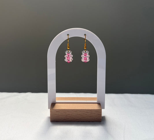 A pair of dangling earrings displayed on a white arched stand with a wooden base. The earrings have gold hooks and clear and pink striped Swarovski crystal  beaded pendants. 