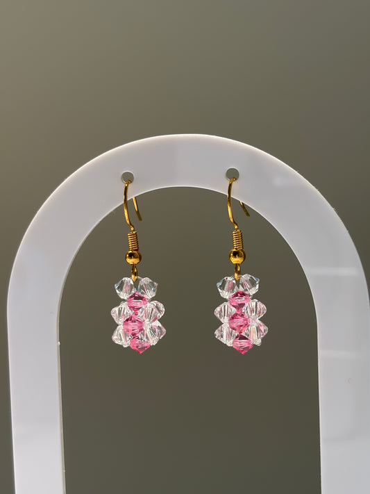A pair of dangling earrings displayed on a white arched stand against a plain gray background. Each earring has a gold-tone fishhook wire and a cluster of faceted clear and pink Swarovski crystal beads at the bottom.