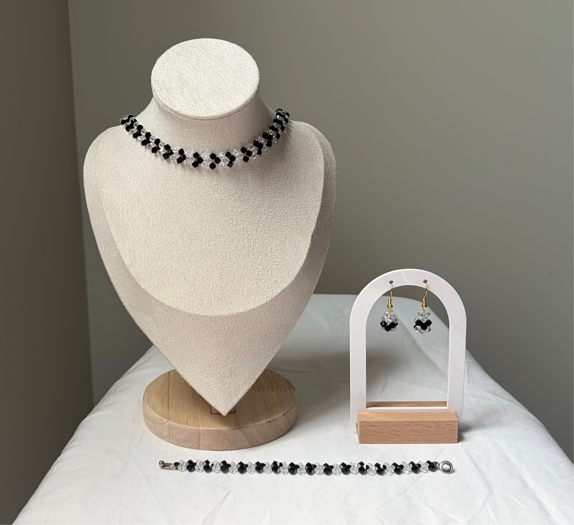 A Swarovski crystal beaded jewelry set displayed on a cream fabric mannequin bust and a small white arch stand, featuring a necklace, bracelet, and matching earrings with black and white v-shaped design. 