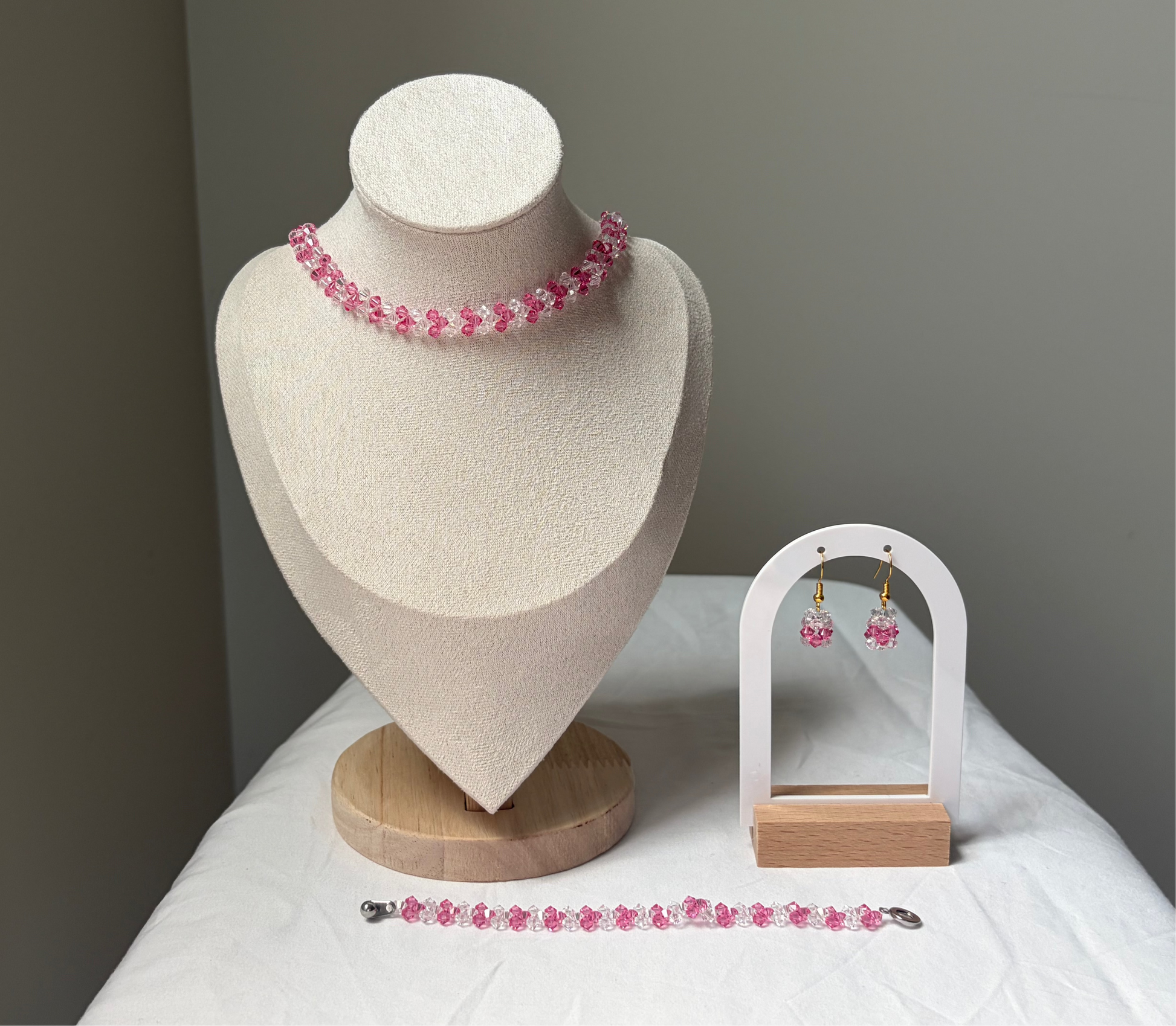 A Swarovski crystal beaded jewelry set displayed on a cream fabric mannequin bust and a small white arch stand, featuring a necklace, bracelet, and matching earrings with pink and white v design. 
