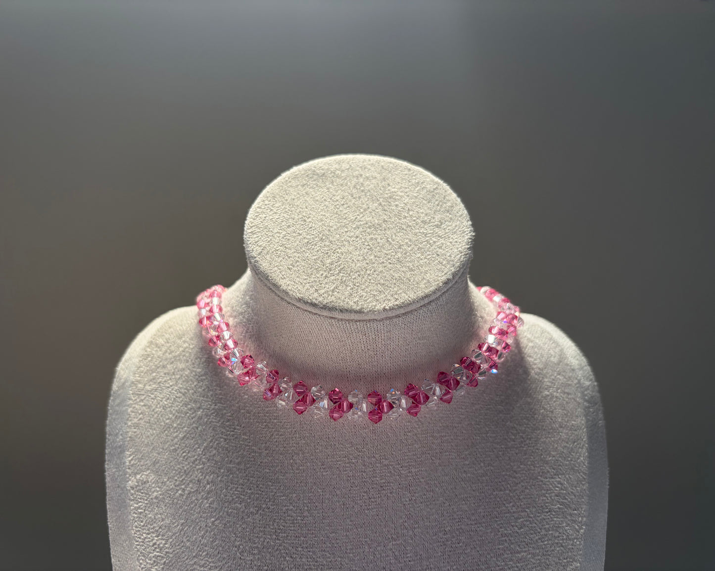 A pink Swarovski crystal beaded v choker necklace displayed on a textured white jewelry bust against a dark gray gradient background.