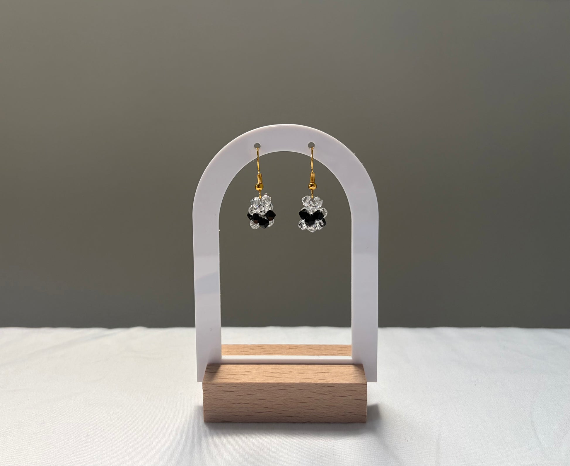 A pair of small dangling earrings displayed on a white arch stand with a wooden base. The earrings have gold hooks and clear faceted Swarovski crystal beads arranged in a black clover-like shape.
