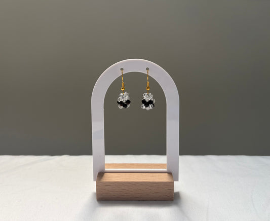 A pair of small dangling earrings displayed on a white arch stand with a wooden base. The earrings have gold hooks and clear faceted Swarovski crystal beads arranged in a black clover-like shape.
