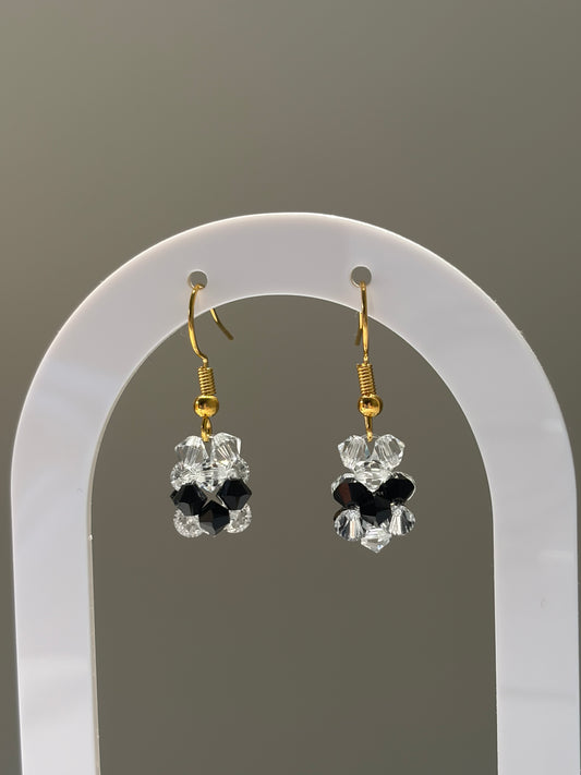 A pair of gold-tone dangle earrings with clear faceted Swarovski crystal beads and small black bead accents, displayed on a white arched stand against a neutral gray background.