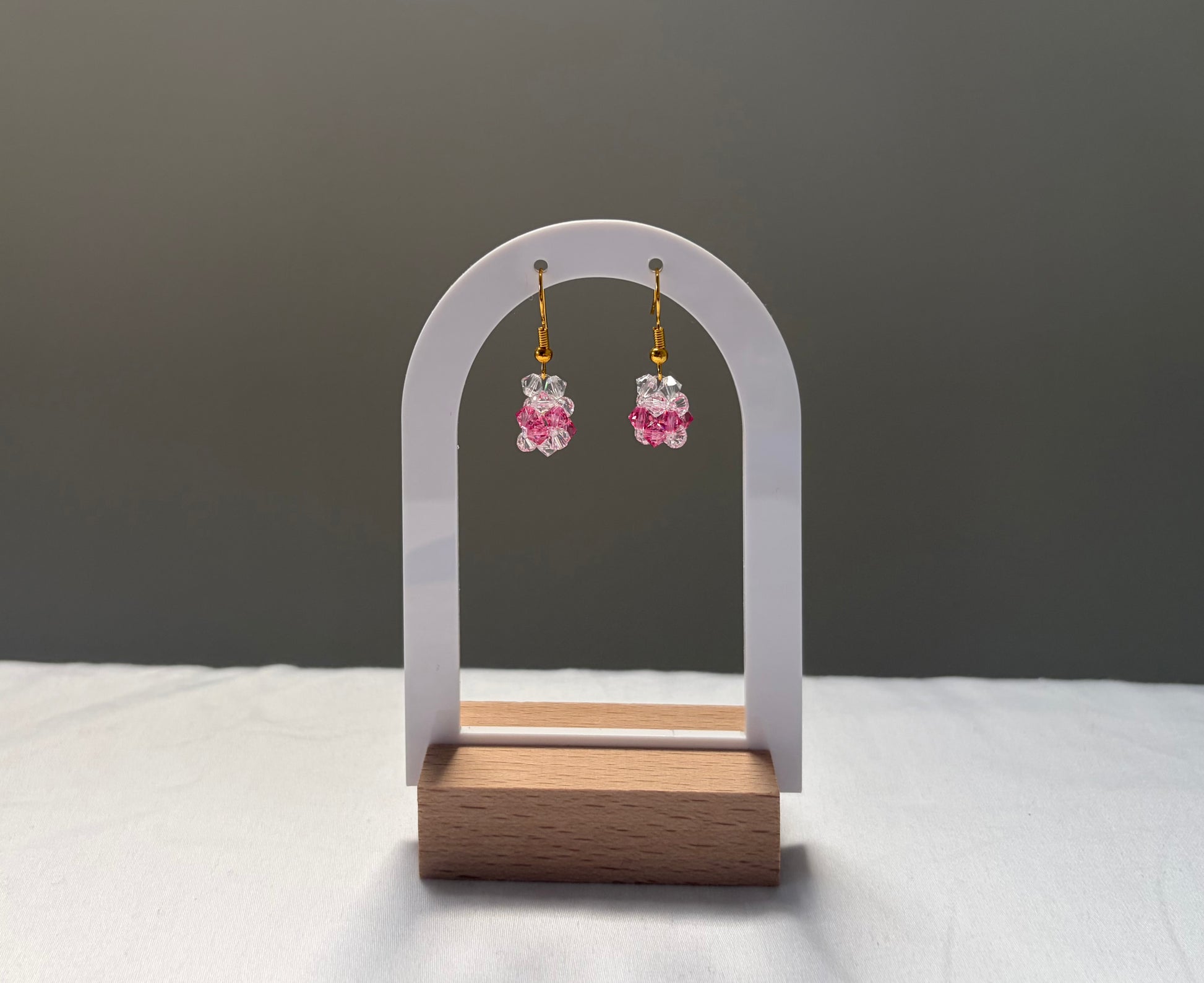 A pair of dangling earrings with pink faceted Swarovski crystal beads and gold hooks, displayed on a white arched stand with a wooden base against a gray background.