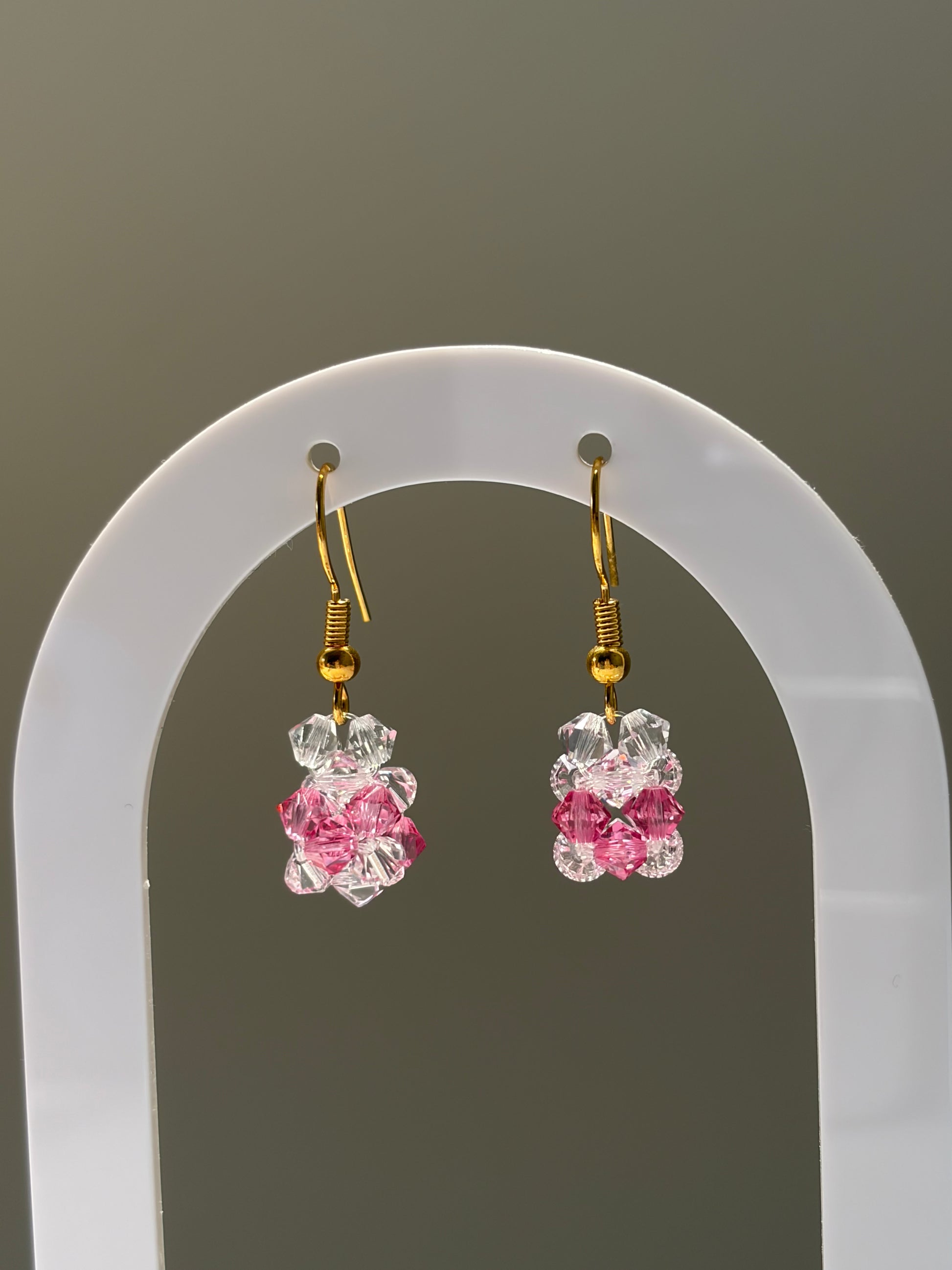 A pair of dangling earrings with gold hooks and clear faceted Swarovski crystal beads transitioning into pink faceted beads, displayed on a white arched stand against a gray background.